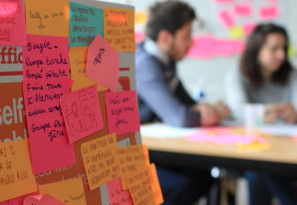 premier plan un taleau rempli de post it, derrière on aperçoit deux personnes assises
