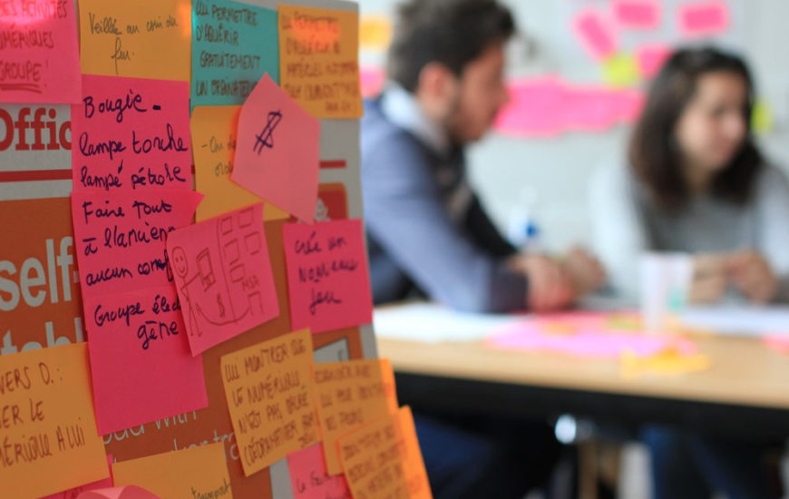 premier plan un taleau rempli de post it, derrière on aperçoit deux personnes assises