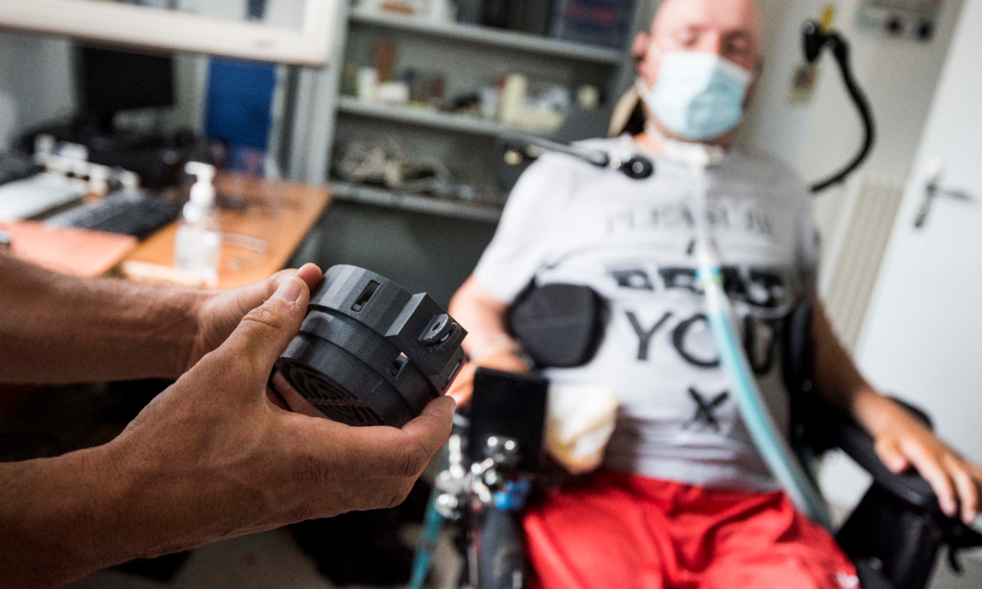 une personne en situation de handicap en arrière plan. Au premier plan une main tenant un objet