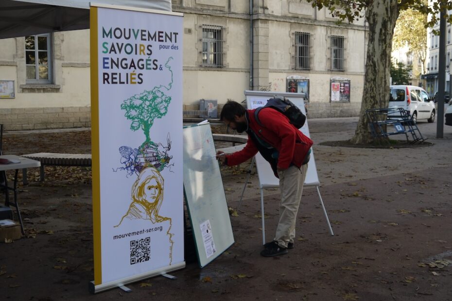 OSER_MSER Un homme écrit sur un tableau devant un stand Mouvement pour des savoirs engagés et reliés, installé en extérieur.