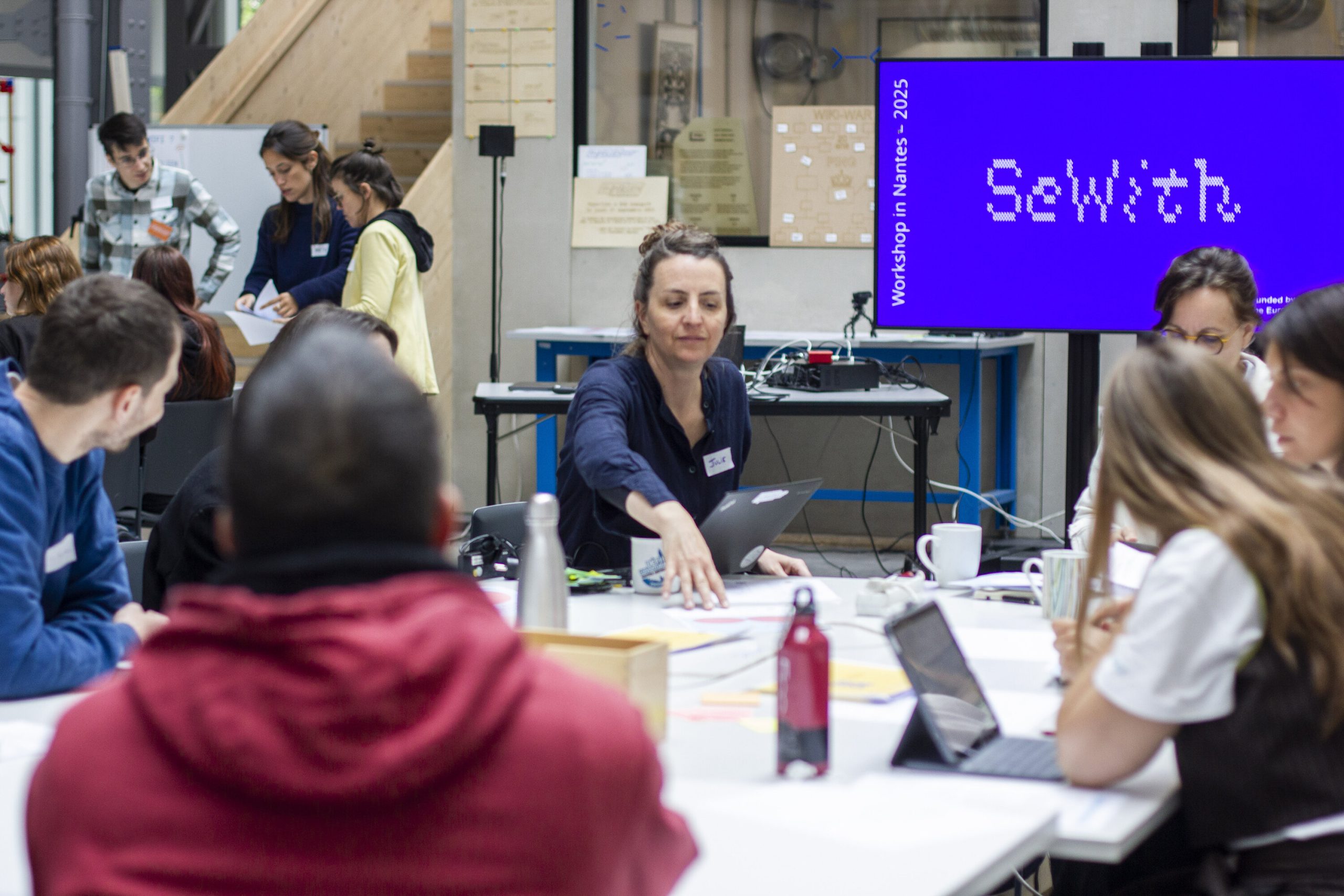 IMG_3613 Une dizaine de personnes dans un fablab assises autour de tables. Dans le fond, on voit un écran avec une présentation titrée "SeWith" sur fond violet
